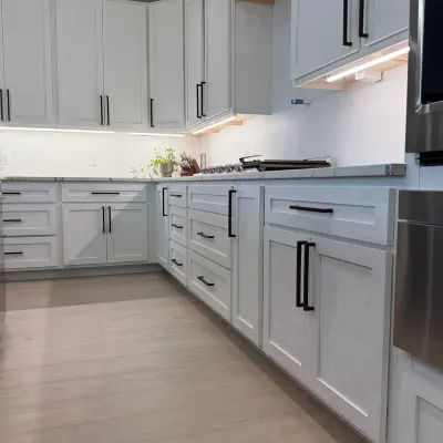 A modern kitchen featuring light grey shaker-style cabinets with black hardware, stainless steel appliances, and light wood plank flooring.
