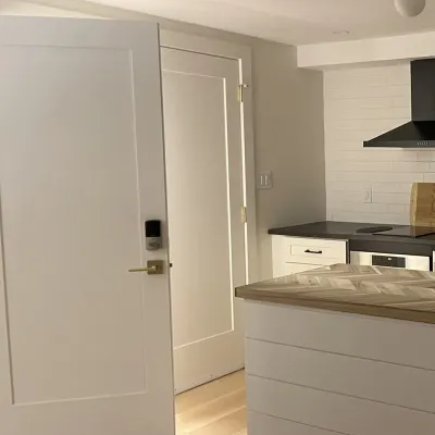A bright kitchen interior featuring light wood flooring, a white interior door with black hardware, and a kitchen island with white shiplap siding and a herringbone wood countertop.