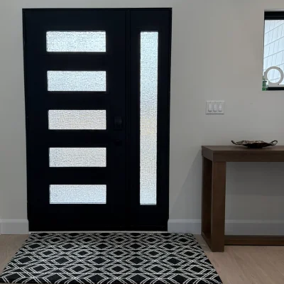 A modern home entryway featuring a black front door with frosted glass inserts, light wood flooring, a black and white geometric rug, and a wooden console table.